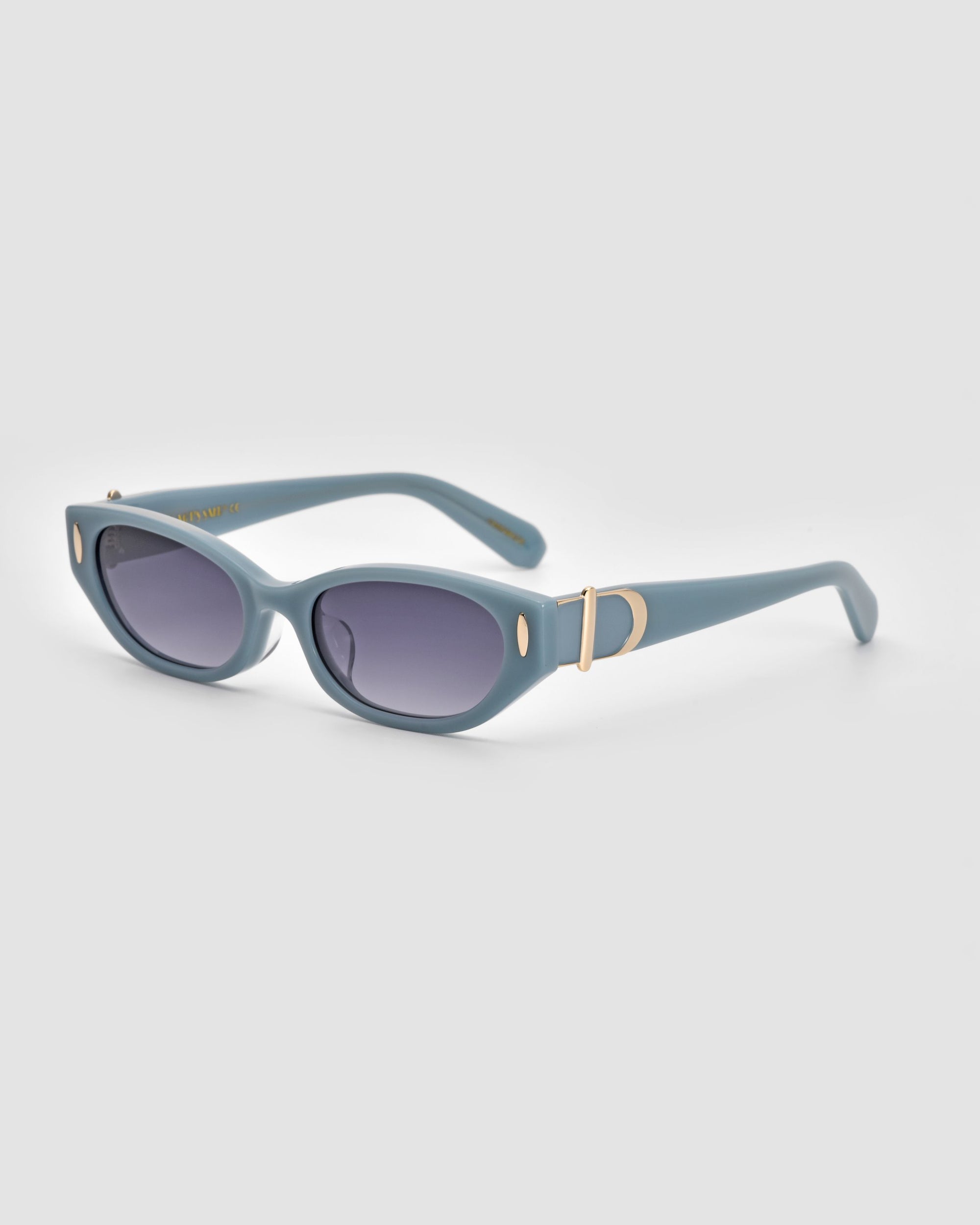 A pair of Skinny Latte cat-eye sunglasses from For Art&#39;s Sake® features light blue frames and dark tinted lenses. The arms are embellished with gold accents, including a small oval cutout near the hinge and a larger oval emblem with a horizontal bar design. Crafted from sustainable materials, these shades exude minimalist elegance against a plain light grey background.