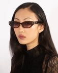 A person with long, straight black hair wearing For Art's Sake® Skinny Latte, small glossy black acetate sunglasses and a black lace top gazes into the distance against a plain white background, exuding retro charm.