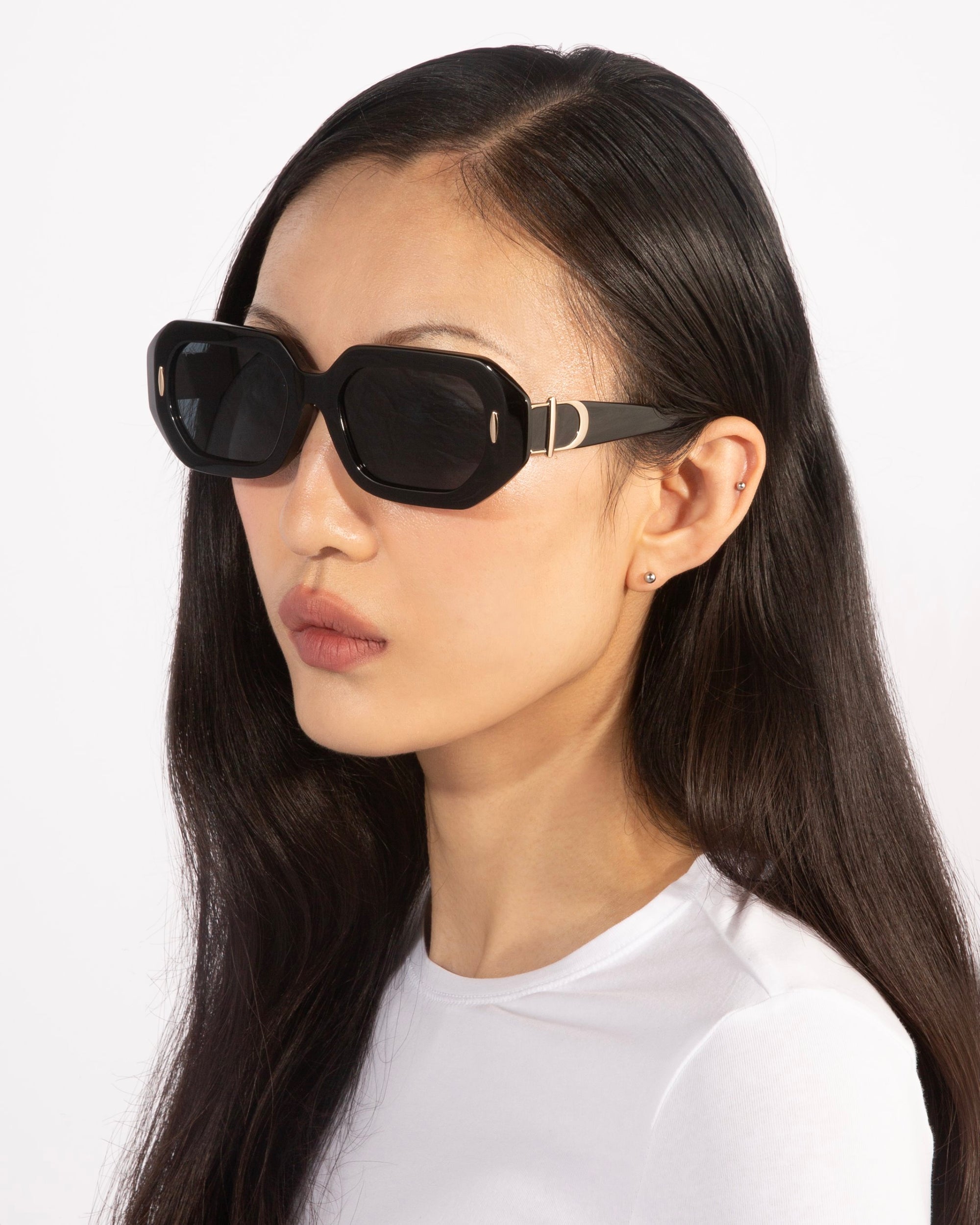 A person with long, sleek dark hair is wearing stylish, oversized black acetate sunglasses from For Art's Sake® and a simple white shirt. The individual has a serious expression and is captured against a plain, light background.