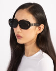 A person with long, sleek dark hair is wearing stylish, oversized black acetate sunglasses from For Art's Sake® and a simple white shirt. The individual has a serious expression and is captured against a plain, light background.