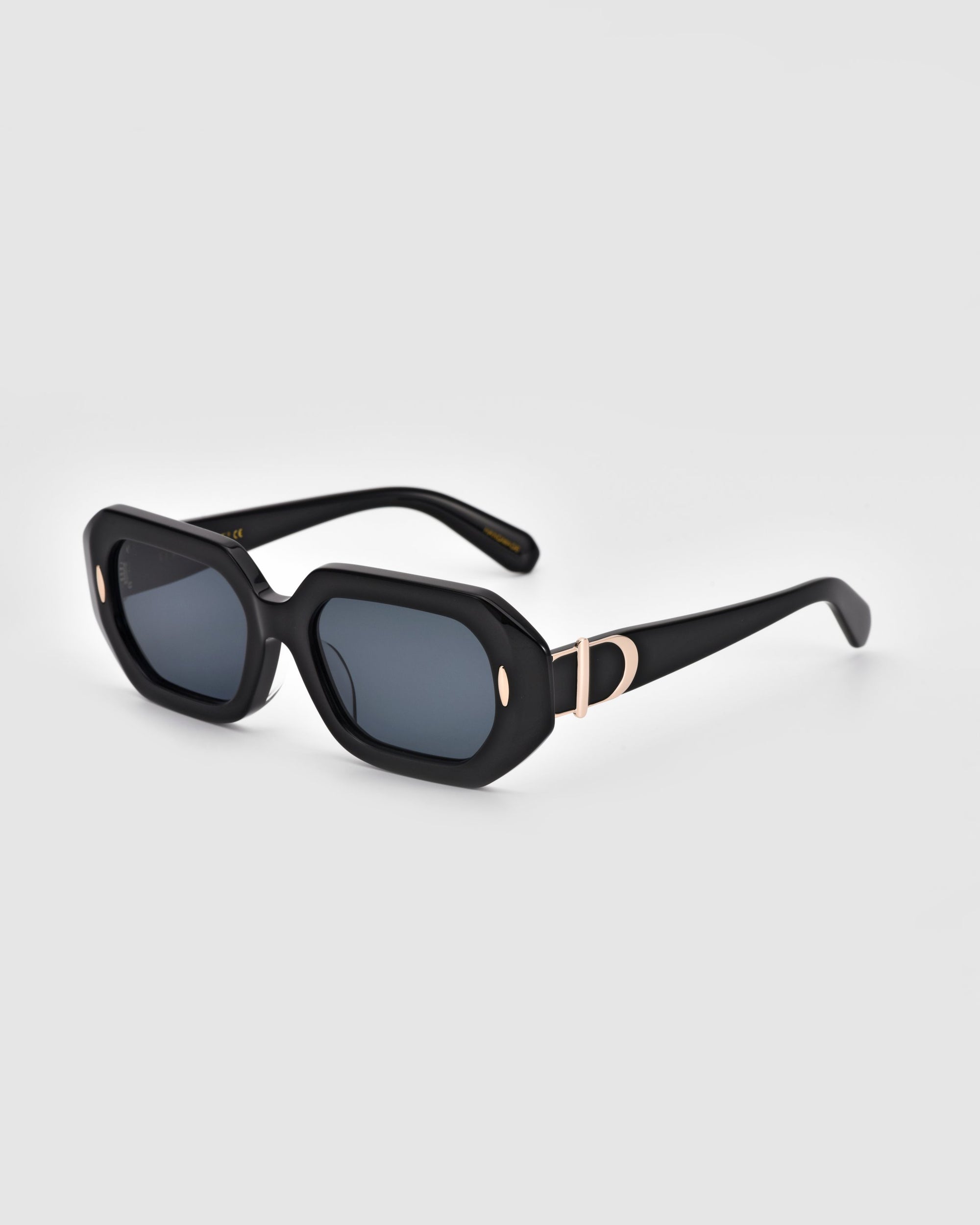 A pair of black rectangular For Art's Sake® Vitamin D sunglasses with dark tinted lenses. The frame features gold oval accents at the corners and a gold "D" detail on the temples near the hinges. Crafted from stainless steel and biodegradable acetate, these sunglasses are set against a plain white background.