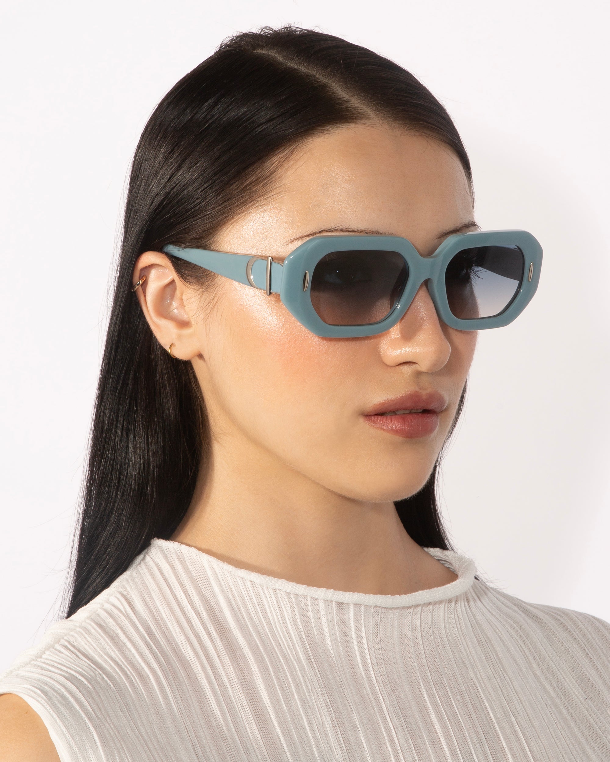 A person with long, straight black hair is wearing blue, hexagonal sunglasses from For Art&#39;s Sake® and a white, sleeveless top. The retro glamour of their look is accentuated by the geometric Vitamin D sunglasses against the plain white background.