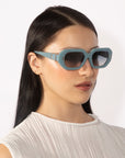 A person with long, straight black hair is wearing blue, hexagonal sunglasses from For Art's Sake® and a white, sleeveless top. The retro glamour of their look is accentuated by the geometric Vitamin D sunglasses against the plain white background.