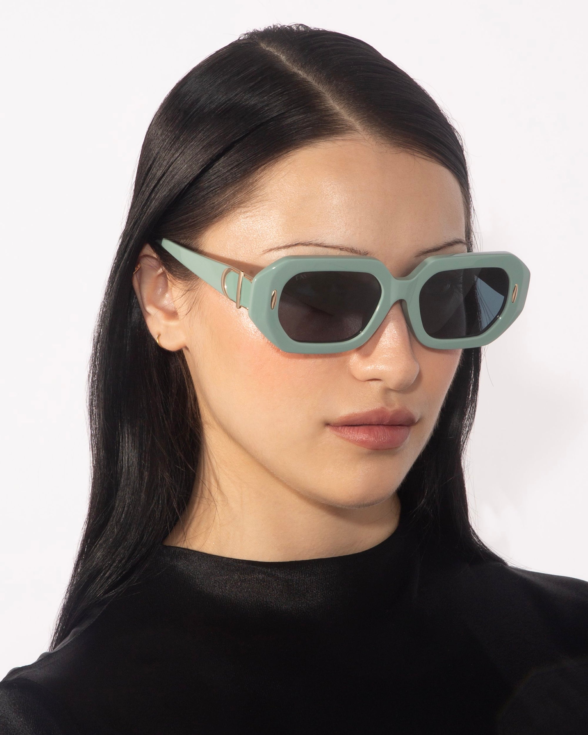 A person with long, straight black hair wears the For Art&#39;s Sake® Vitamin D oversized, rectangular mint green geometric sunglasses and a black top. The background is white. They are gazing slightly to the side with a neutral expression, exuding retro glamour.