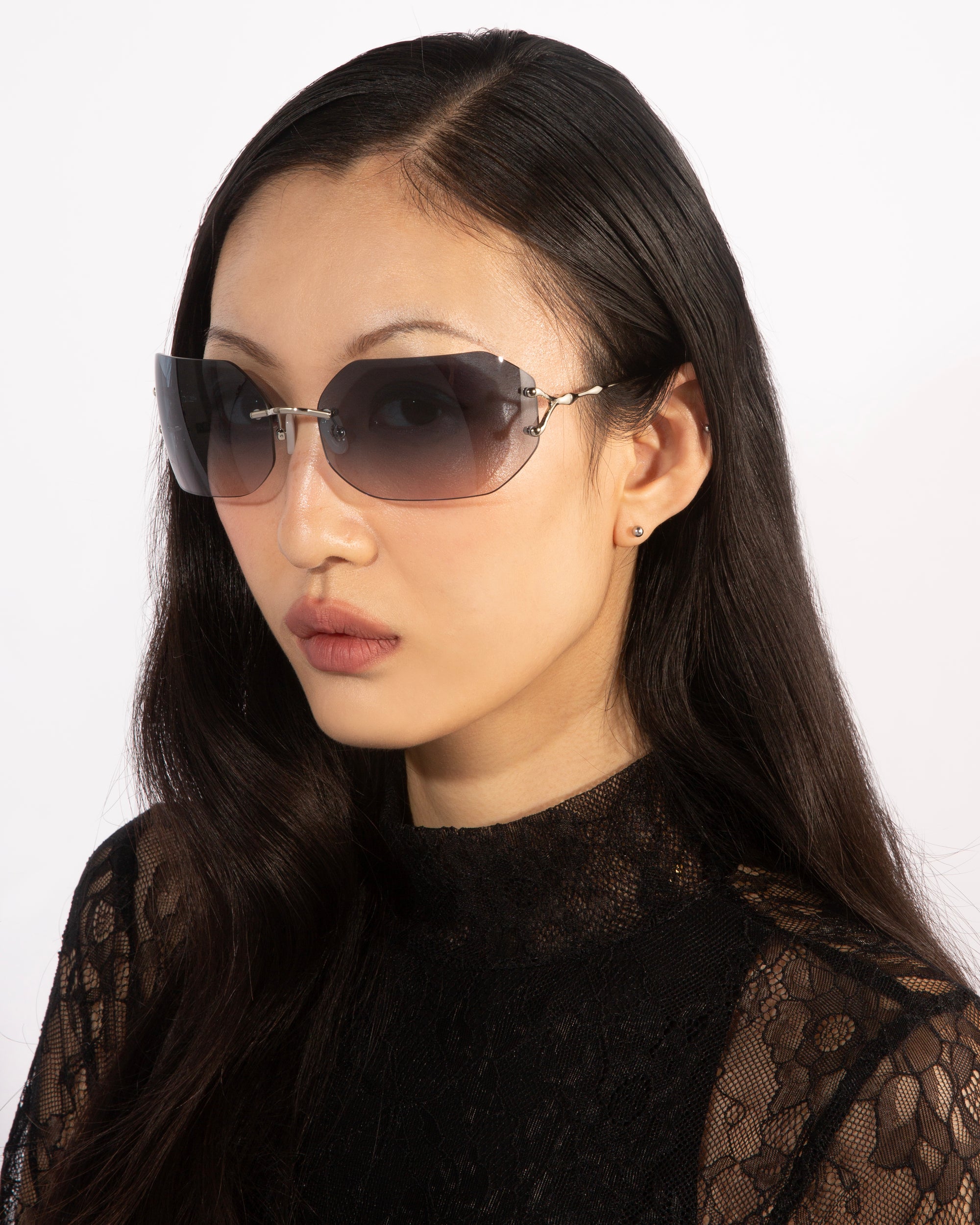 A person with long dark hair wearing large, stylish For Art's Sake® Hutton sunglasses and a black lace top exudes vintage glamour as they look off to the side against a plain white background.