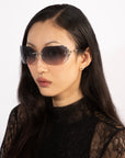 A person with long dark hair wearing large, stylish For Art's Sake® Hutton sunglasses and a black lace top exudes vintage glamour as they look off to the side against a plain white background.