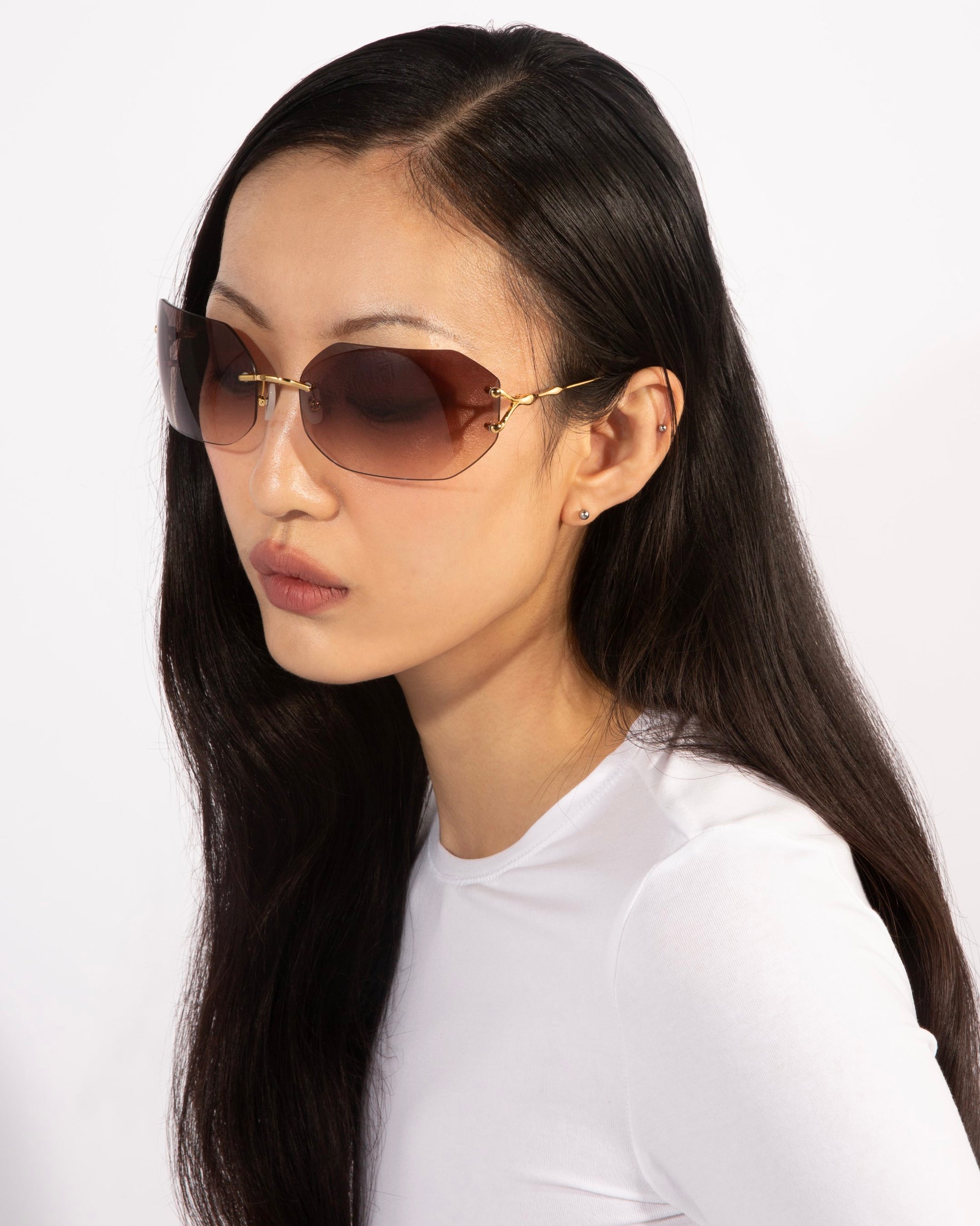 A person with long dark hair is wearing oversized, rimless Hutton sunglasses from For Art&#39;s Sake® featuring gradient brown lenses and a white long-sleeve shirt, exuding vintage glamour. The background is a plain, light color.