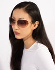 A person with long dark hair is wearing oversized, rimless Hutton sunglasses from For Art's Sake® featuring gradient brown lenses and a white long-sleeve shirt, exuding vintage glamour. The background is a plain, light color.