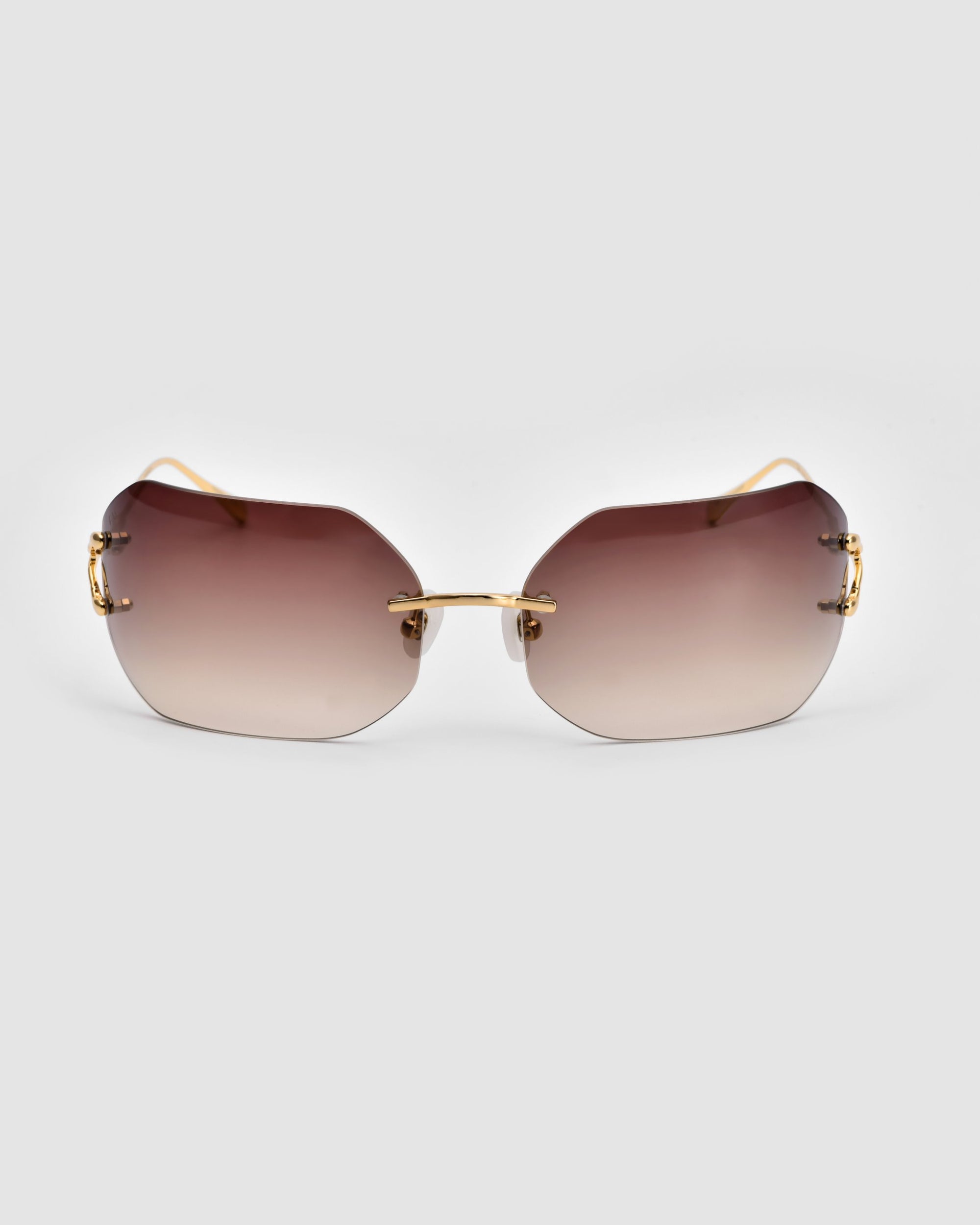 A pair of Hutton sunglasses by For Art&#39;s Sake®, featuring brown gradient lenses and gold-tone metal accents, set against a plain gray background. These sunglasses exude vintage glamour with their frameless design and sleek, curved arms.