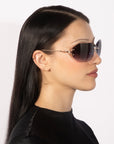 A person with long black hair is shown in profile wearing the large, dark Hutton sunglasses from For Art's Sake®. They are dressed in a black top and have small, subtle earrings that add a touch of vintage glamour against the plain white background.