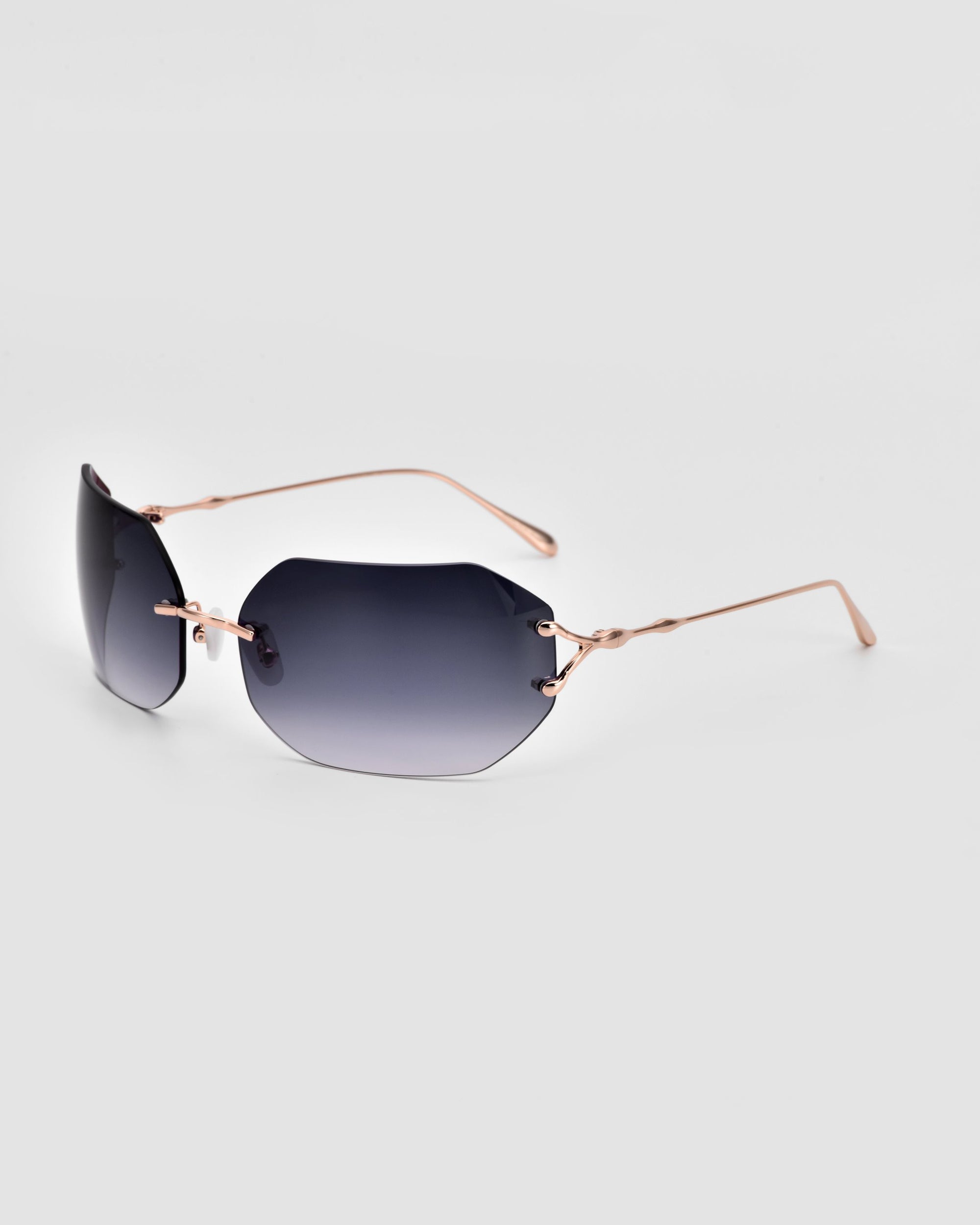 A pair of For Art's Sake® Hutton sunglasses with dark blue lenses and thin, rose gold temples is displayed against a plain white background. The frameless design gives these sunglasses an iconic style, boasting a sleek and modern look.