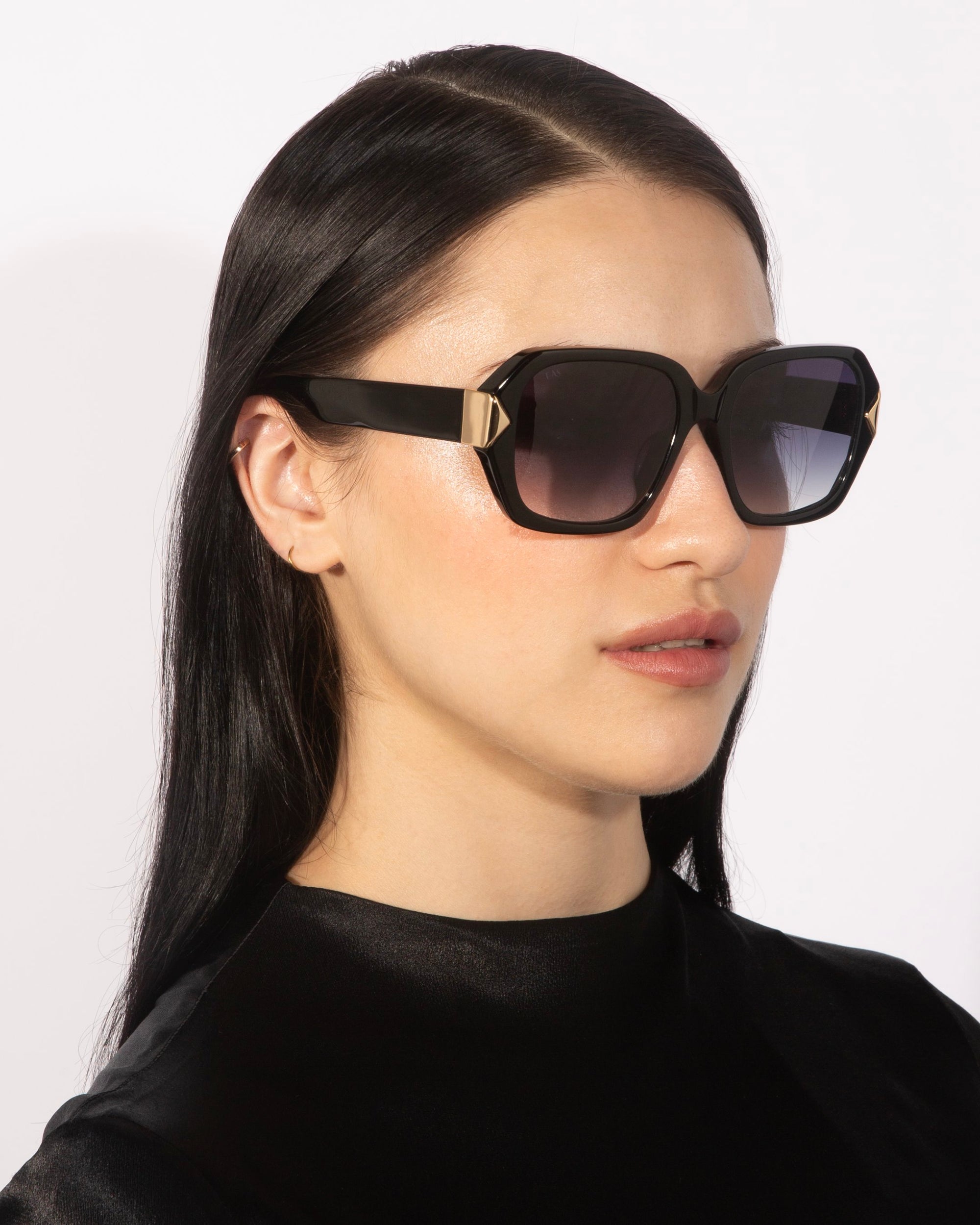 A person with long, black hair wears the Frankie oversized geometric sunglasses from For Art&#39;s Sake®, featuring chunky acetate frames and gold accents. They are dressed in a black, shiny top and posed against a light background.