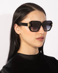 A person with long, black hair wears the Frankie oversized geometric sunglasses from For Art's Sake®, featuring chunky acetate frames and gold accents. They are dressed in a black, shiny top and posed against a light background.