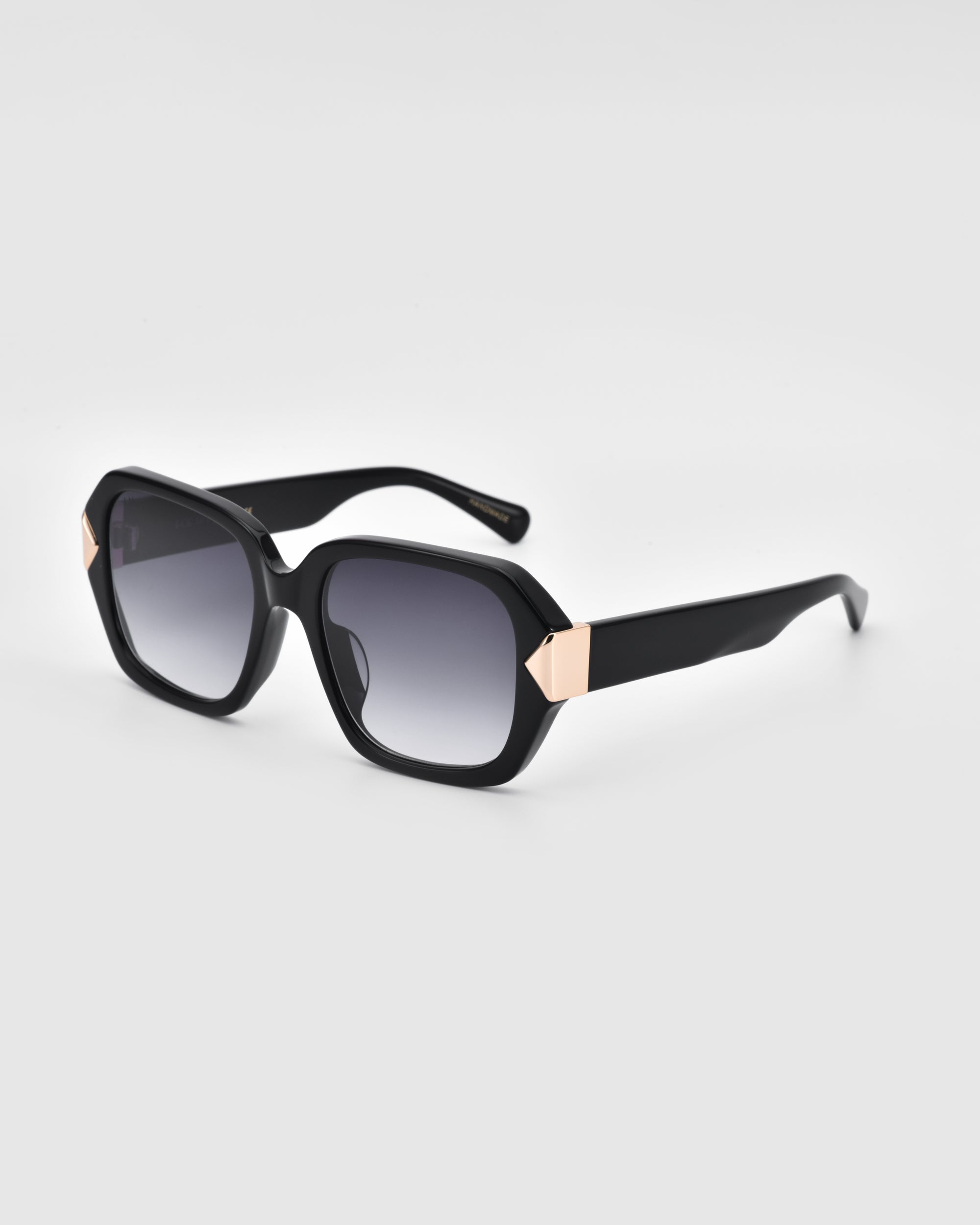 A pair of stylish, rectangular Frankie sunglasses by For Art&#39;s Sake®, featuring black frames with gradient dark lenses. These timeless sunglasses have thick black frames adorned with small gold triangular details at the corners near the lenses, all set against a clean, light grey background.