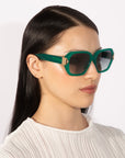 A person with long, straight black hair wears the Frankie oversized geometric sunglasses by For Art's Sake®, featuring a metallic accent on the temples. They have a neutral expression and are dressed in a light-colored, textured top, photographed against a plain white background.