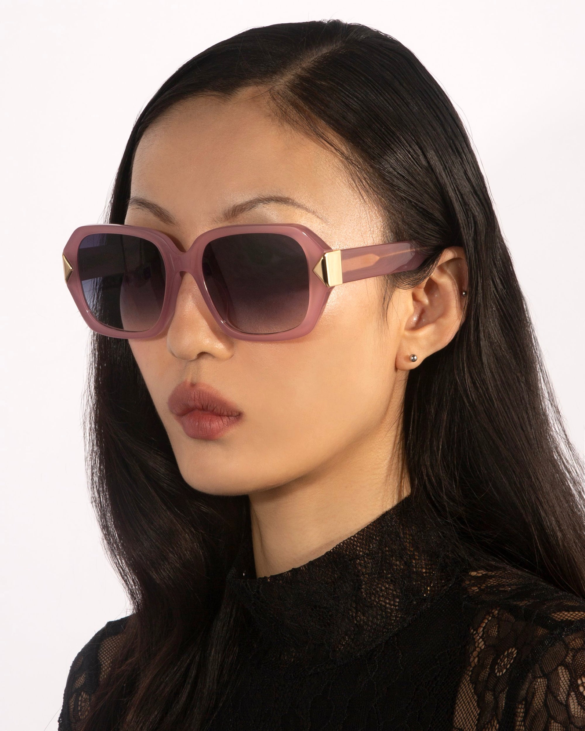 A woman with long, dark hair is wearing oversized geometric Frankie sunglasses by For Art&#39;s Sake®, featuring gradient lenses and gold accents. She has a small earring and is dressed in a black lace top, looking straight ahead against a plain background.