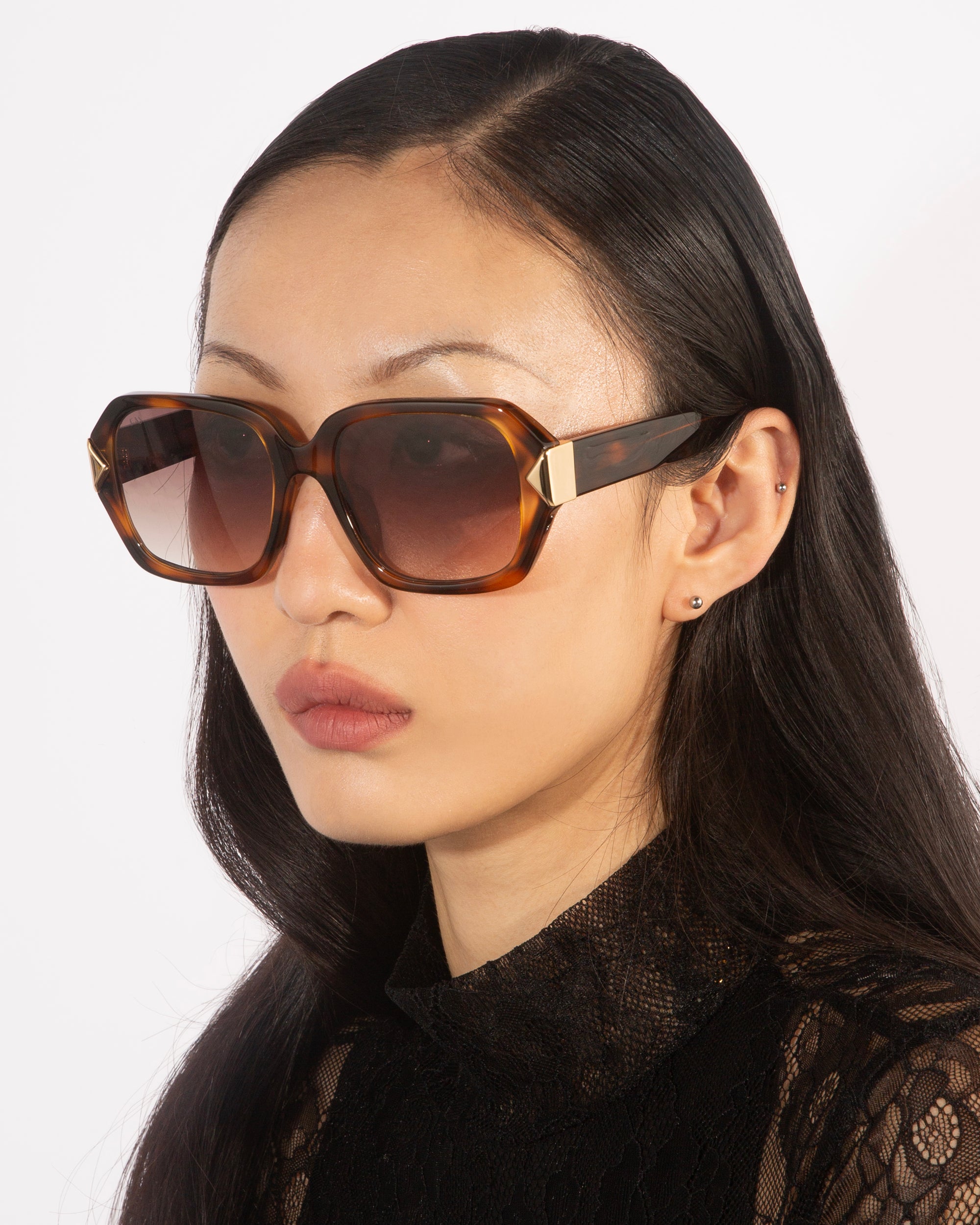 A woman with long dark hair wears oversized geometric Frankie sunglasses with gradient lenses by For Art's Sake®, paired with a black lace top. She is photographed from the shoulders up, against a plain white background, looking to the left with a neutral expression.