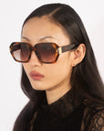 A woman with long dark hair wears oversized geometric Frankie sunglasses with gradient lenses by For Art's Sake®, paired with a black lace top. She is photographed from the shoulders up, against a plain white background, looking to the left with a neutral expression.