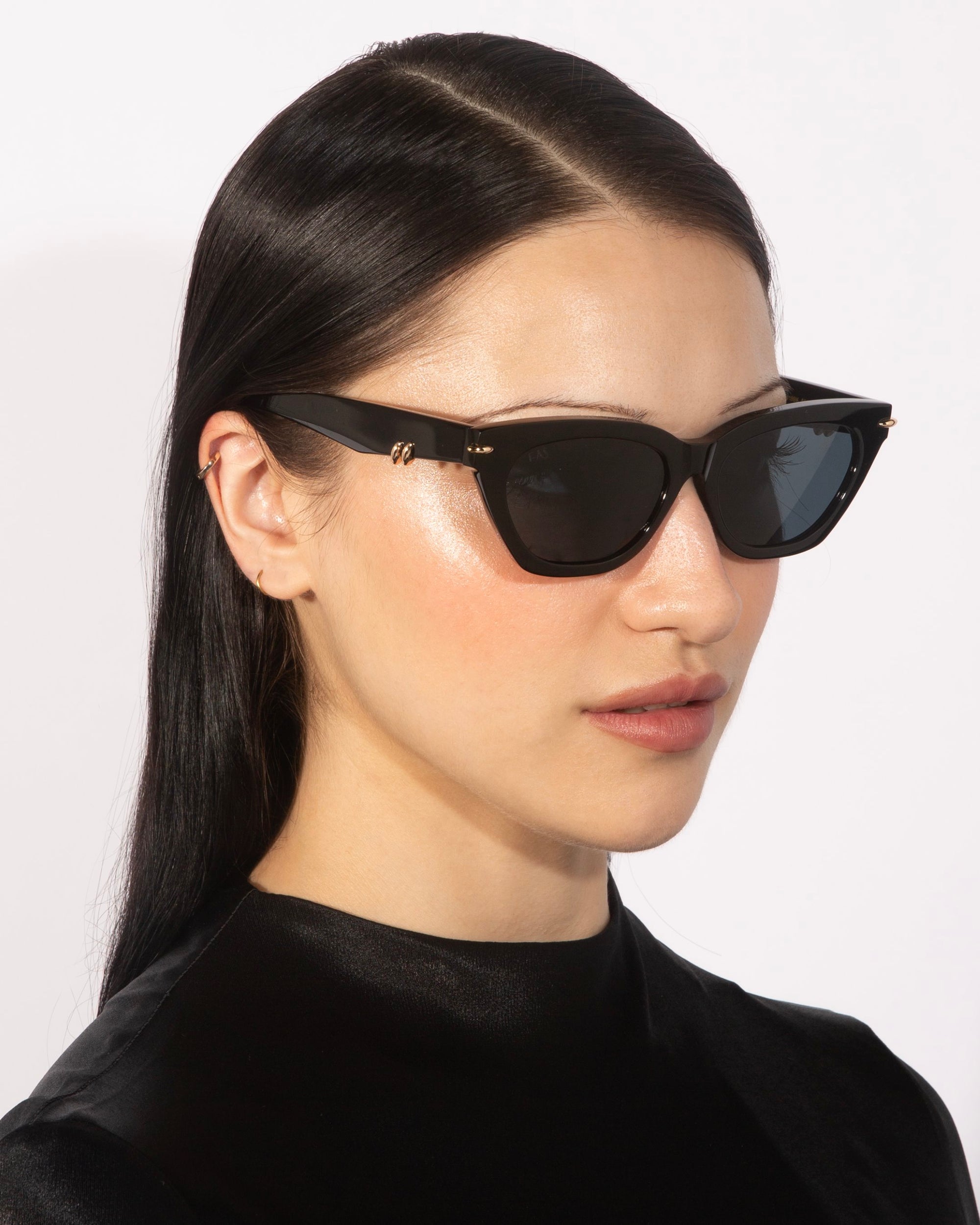 A person wearing For Art&#39;s Sake® Bonbon black rectangular sunglasses adorned with handcrafted metal decorations and a black jacket, glances slightly to the side. Their long dark hair is pulled back, and they are posed against a plain white background.