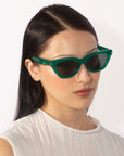 A person with long black hair is wearing For Art's Sake® Bonbon sunglasses, which feature a cat-eye silhouette with green frames and tinted lenses. They have a neutral expression and are dressed in a white textured shirt against a plain, light background.