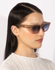 A person with straight black hair wearing stylish For Art's Sake® Bonbon sunglasses in a cat-eye silhouette and a white textured top. The individual is looking to the right and has minimal makeup. The background is plain and well-lit.