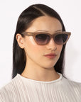 A person with long, straight black hair is wearing a pair of stylish Bonbon sunglasses from For Art's Sake®, featuring gradient lenses. They are dressed in a light-colored, textured top with a high neckline and are posing against a plain white background.