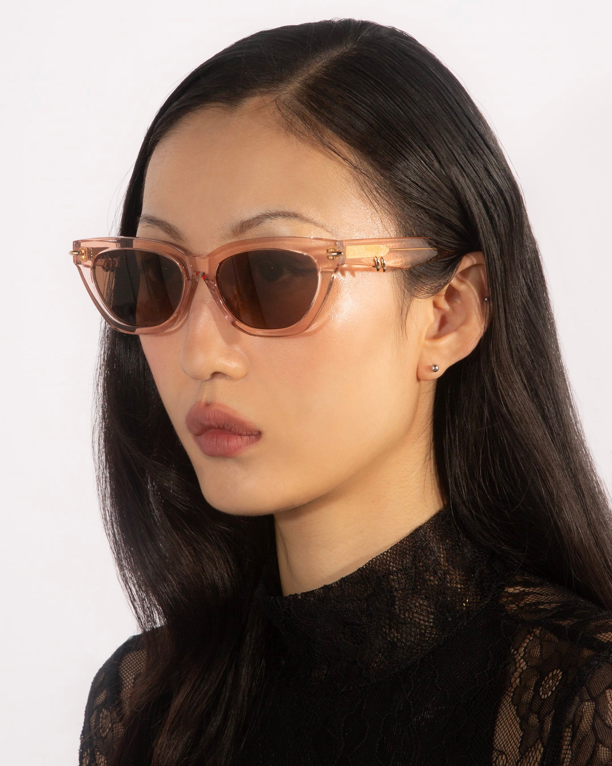 A woman with long black hair dons a pair of For Art&#39;s Sake® Bonbon sunglasses in pink translucent with a cat-eye silhouette, complemented by a black lace high-neck top. She gazes away from the camera against a plain white background, emphasizing her fashionable and poised demeanor.