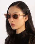 A woman with long black hair dons a pair of For Art's Sake® Bonbon sunglasses in pink translucent with a cat-eye silhouette, complemented by a black lace high-neck top. She gazes away from the camera against a plain white background, emphasizing her fashionable and poised demeanor.