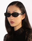 A person with long dark hair is wearing For Art's Sake® Friday geometric eyewear with UV protection and a black lace top. The background is a plain white.