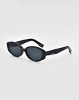 A pair of For Art's Sake® Friday sunglasses with black oval-shaped frames and dark lenses is displayed against a plain white background. These stylish sunglasses feature thick black frames, UV protection, and a small silver embellishment on the temple arms.