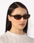 A woman with straight, long black hair wears For Art's Sake® Friday rectangular tortoiseshell sunglasses, which offer UV protection. She has a neutral expression and is dressed in a white, textured blouse. The background is plain and well-lit, highlighting her and the geometric eyewear.