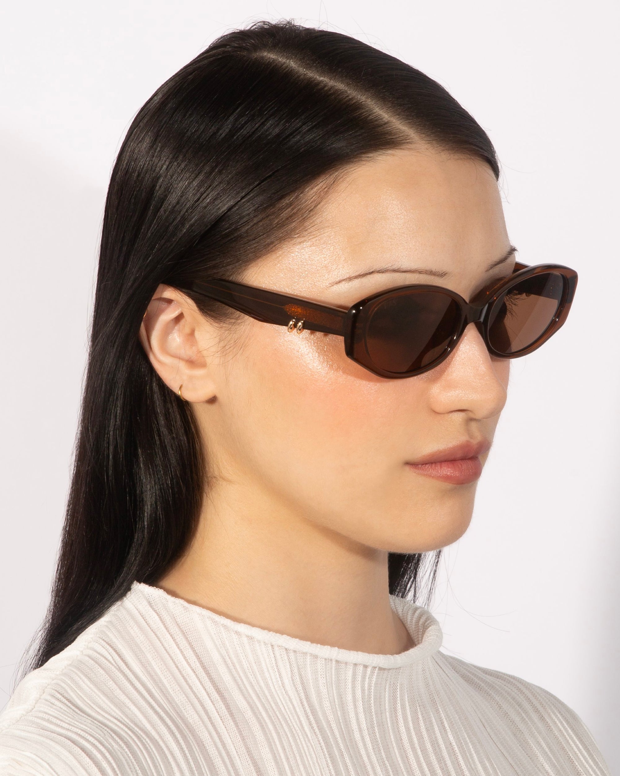 A person with long, dark hair wearing For Art's Sake® Friday tortoiseshell rectangular sunglasses with UV protection and a white textured top is looking to the side against a plain background.