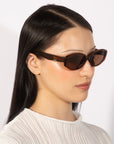 A person with long, dark hair wearing For Art's Sake® Friday tortoiseshell rectangular sunglasses with UV protection and a white textured top is looking to the side against a plain background.