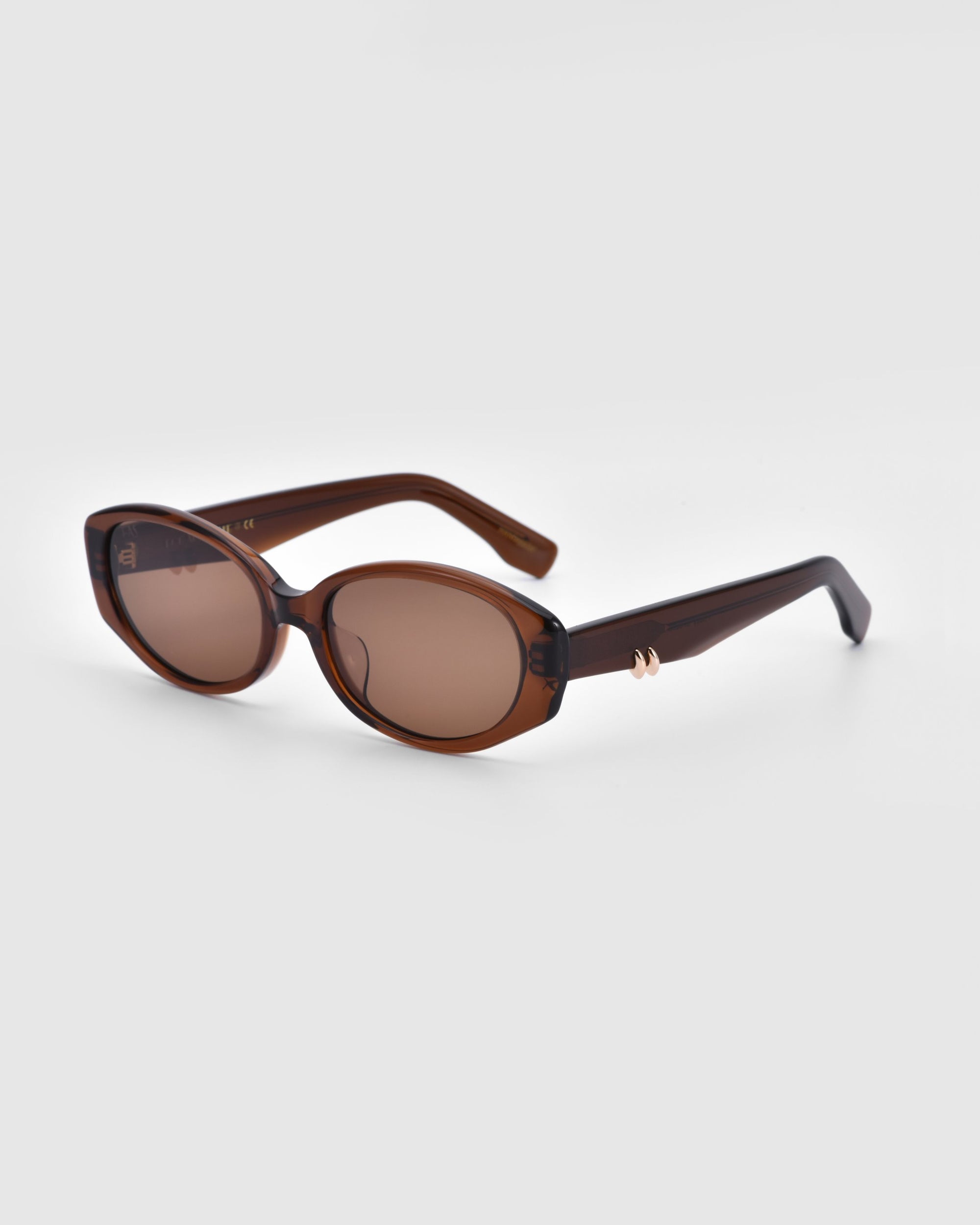 A pair of brown sunglasses from For Art's Sake® with oval-shaped lenses and dark brown frames. The Friday sunglasses have sleek, slightly curved arms adorned with a small golden accent near the hinges. Featuring UV protection and tinted brown lenses, these stylish shades stand out against the plain white background.