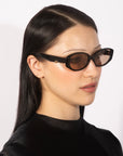 A person with long, straight black hair wears stylish Friday sunglasses by For Art's Sake® featuring dark lenses that provide UV protection. They have a helix piercing on their ear and are dressed in a black top against a plain white background.