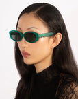 A person with long, straight black hair dons a pair of For Art's Sake® Friday green oval sunglasses, which provide UV protection, along with a black lace top. They sport multiple small earrings in their left ear and gaze to the left with a neutral expression against a plain white background.