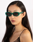 A person with long black hair wearing For Art's Sake® Friday green rectangular sunglasses and a black lace top gazes to the side against a plain white background. With small earrings and UV protection geometric eyewear, they have a calm expression.