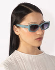 A person with black hair is wearing the Dyer teal rectangular luxury sunglasses with dark lenses from For Art's Sake®, a white pleated top, and small stud earrings. The background is plain white.