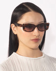 A person with straight, long dark hair is wearing the Dyer rectangular brown sunglasses by For Art's Sake® and a white textured top against a luxurious plain white background.