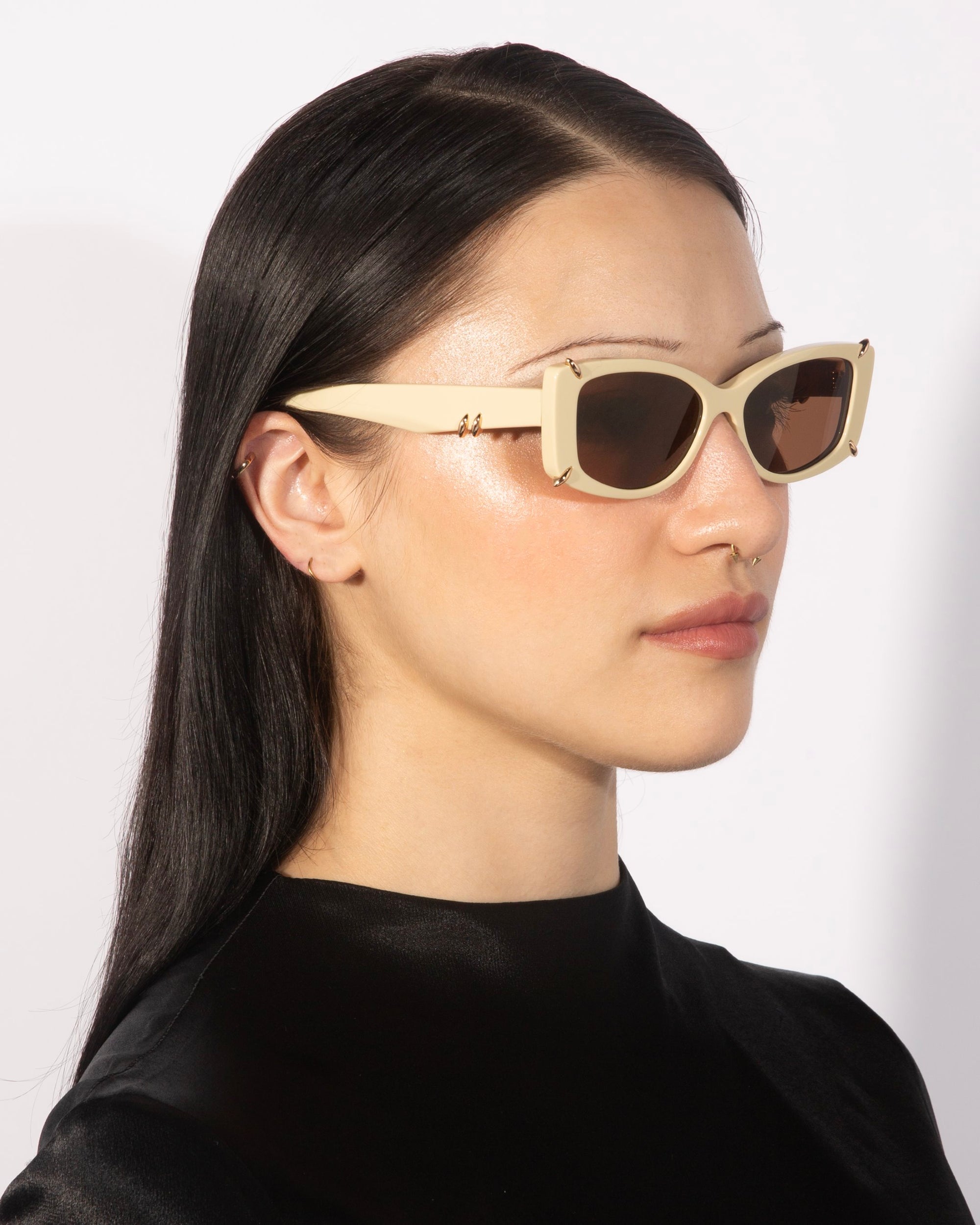 A person with long, straight dark hair is wearing the luxurious Dyer rectangular sunglasses in a cream color with black lenses from For Art's Sake®, along with a small nose piercing and a black high-neck top. The background is plain white.