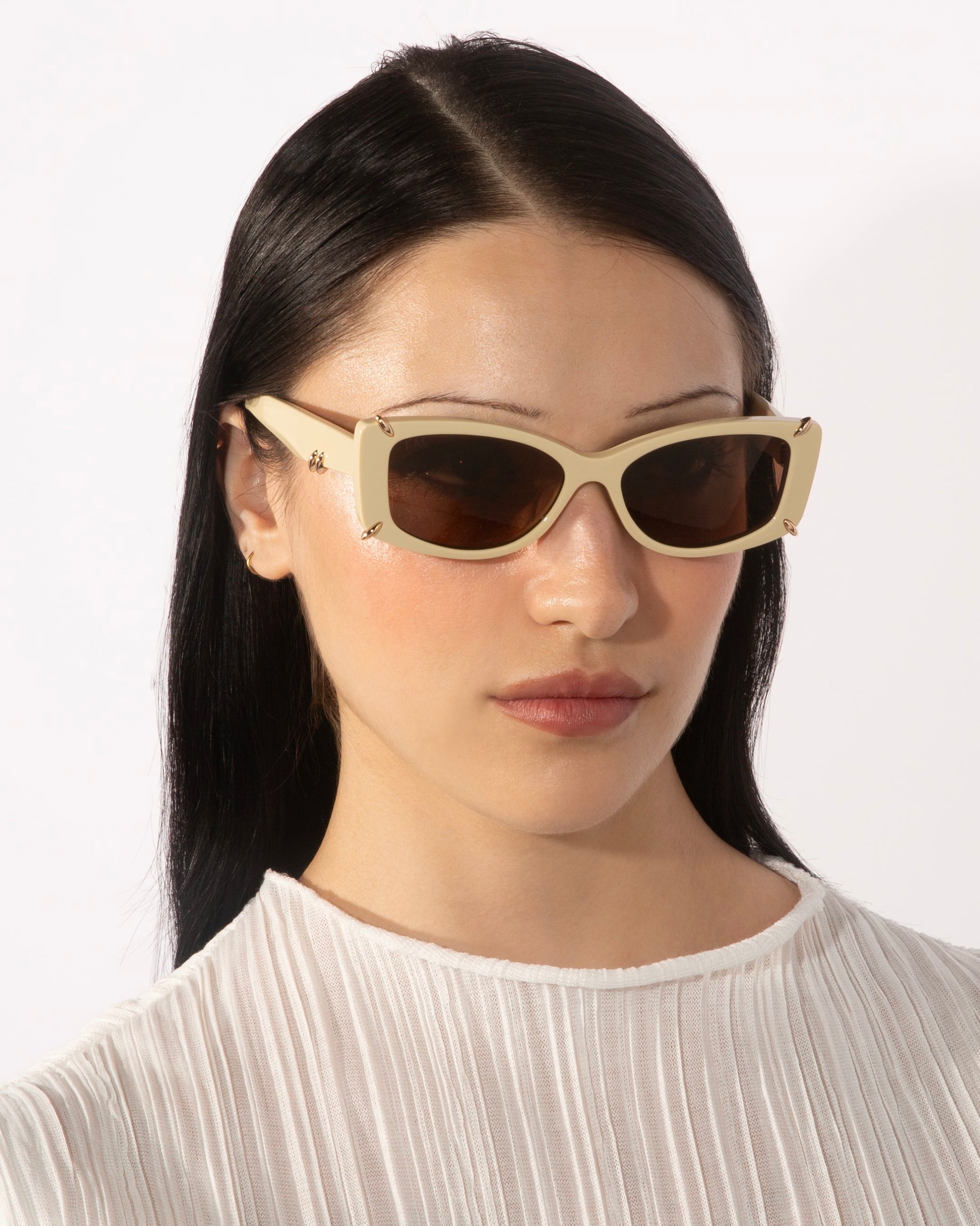 A person with long black hair wears For Art&#39;s Sake® Dyer rectangular beige luxury sunglasses with dark lenses and a white, pleated blouse. The background is plain white.