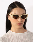 A person with long black hair wears For Art's Sake® Dyer rectangular beige luxury sunglasses with dark lenses and a white, pleated blouse. The background is plain white.