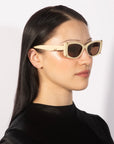 A person with long, straight dark hair is wearing the luxurious Dyer rectangular sunglasses in a cream color with black lenses from For Art's Sake®, along with a small nose piercing and a black high-neck top. The background is plain white.