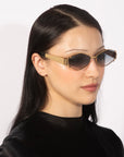 A person with straight black hair dons the narrow, angular For Art's Sake® Empress sunglasses featuring a gold frame and dark lenses. They are outfitted in a black, high-collared ensemble and posed against a plain white background.