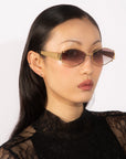 A person with long dark hair is wearing For Art's Sake® Empress sunglasses with gold frames and a black lace top. The photo is taken against a plain white background. The person has a neutral expression and an elegant, minimalist style, showcasing their taste in luxury eyewear.