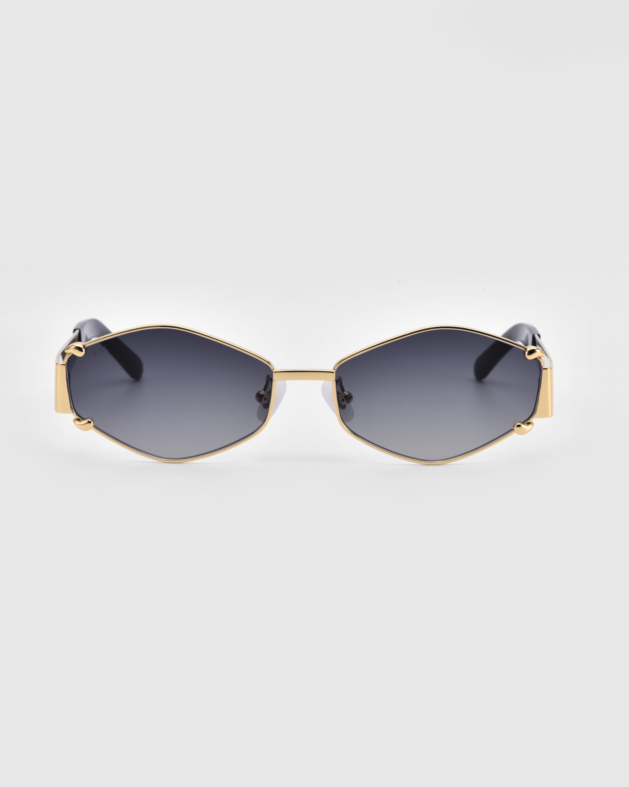 The Empress by For Art&#39;s Sake® are luxurious hexagonal sunglasses featuring black-tinted lenses and a thin gold metal frame. Their oversized design offers UV protection, while the black temples adorned with gold accents add an elegant touch. The background is plain and light-colored.