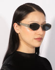 A person with long, straight black hair is wearing fashionable For Art's Sake® Empress sunglasses with narrow rectangular metallic frames. They are dressed in a sleek black top and standing against a plain, light background. Their expression is calm and neutral.