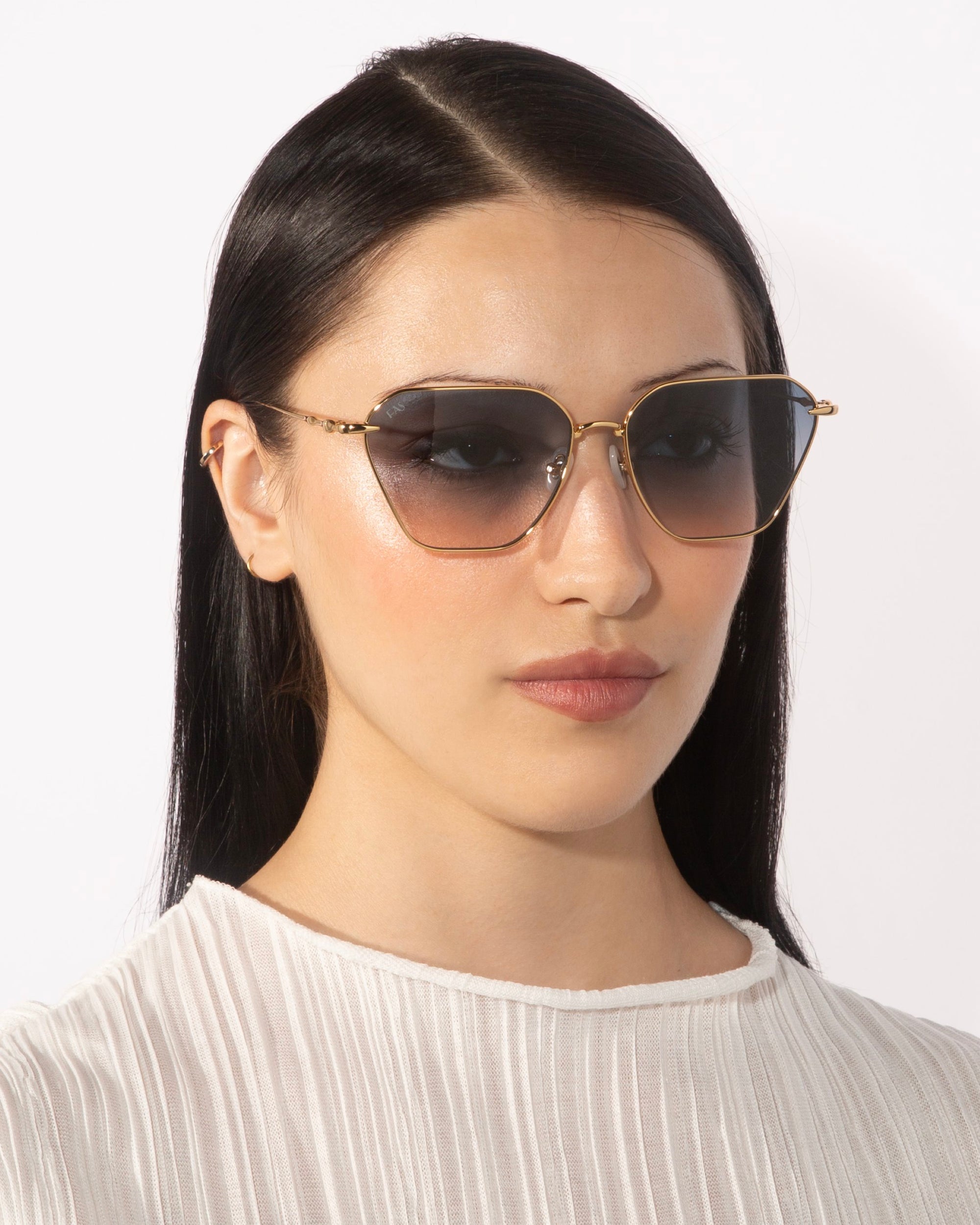 A person with long, straight, dark hair wearing For Art's Sake® Cece sunglasses with gradient lenses and a pleated white blouse stares straight ahead against a plain white background.