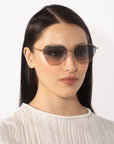 A person with long, straight, dark hair wearing For Art's Sake® Cece sunglasses with gradient lenses and a pleated white blouse stares straight ahead against a plain white background.