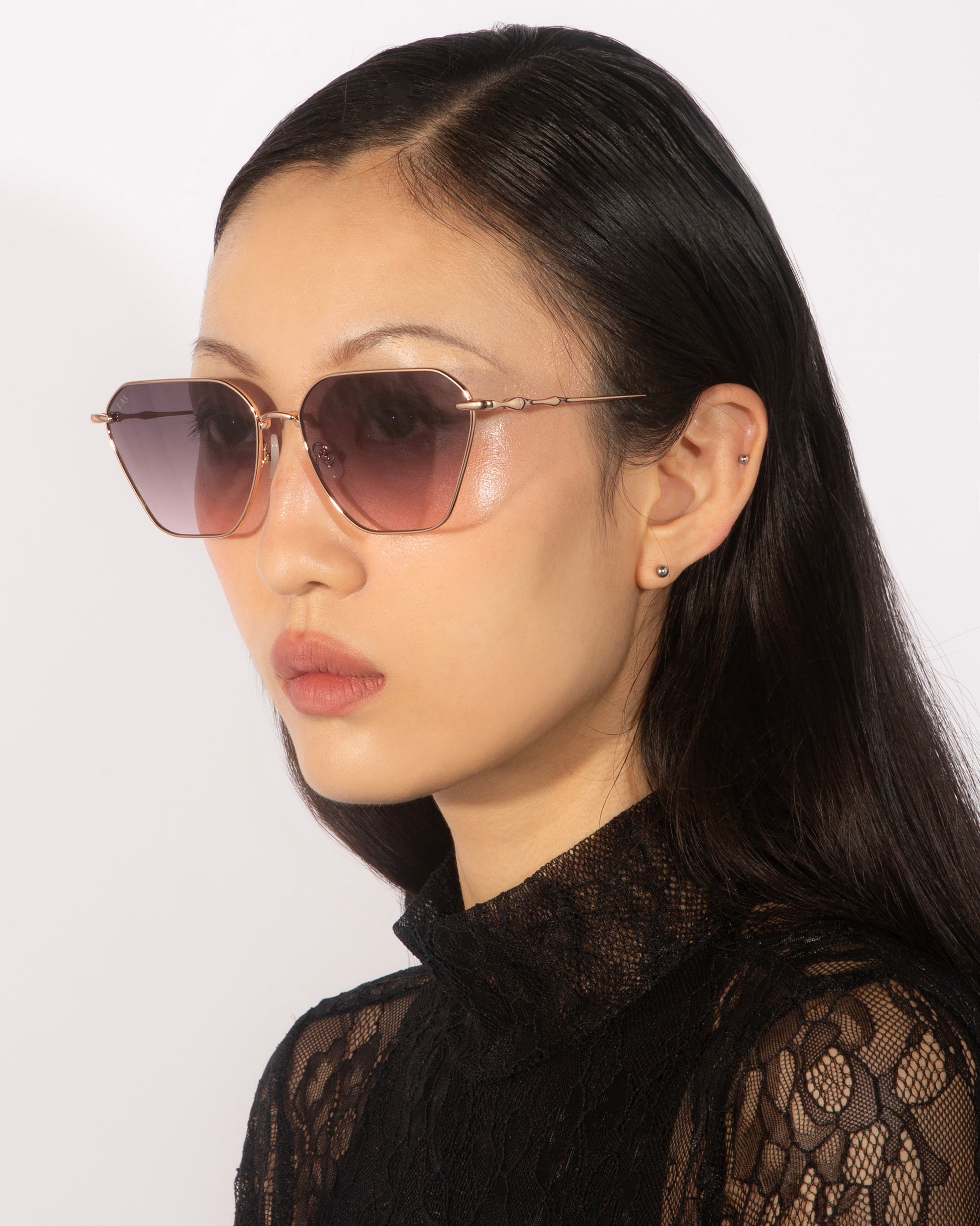 A person with long dark hair is wearing stylish Cece sunglasses from For Art's Sake®, featuring gradient lenses and a hexagonal frame. They are also wearing a black lace top and small earrings. The background is plain and neutral.